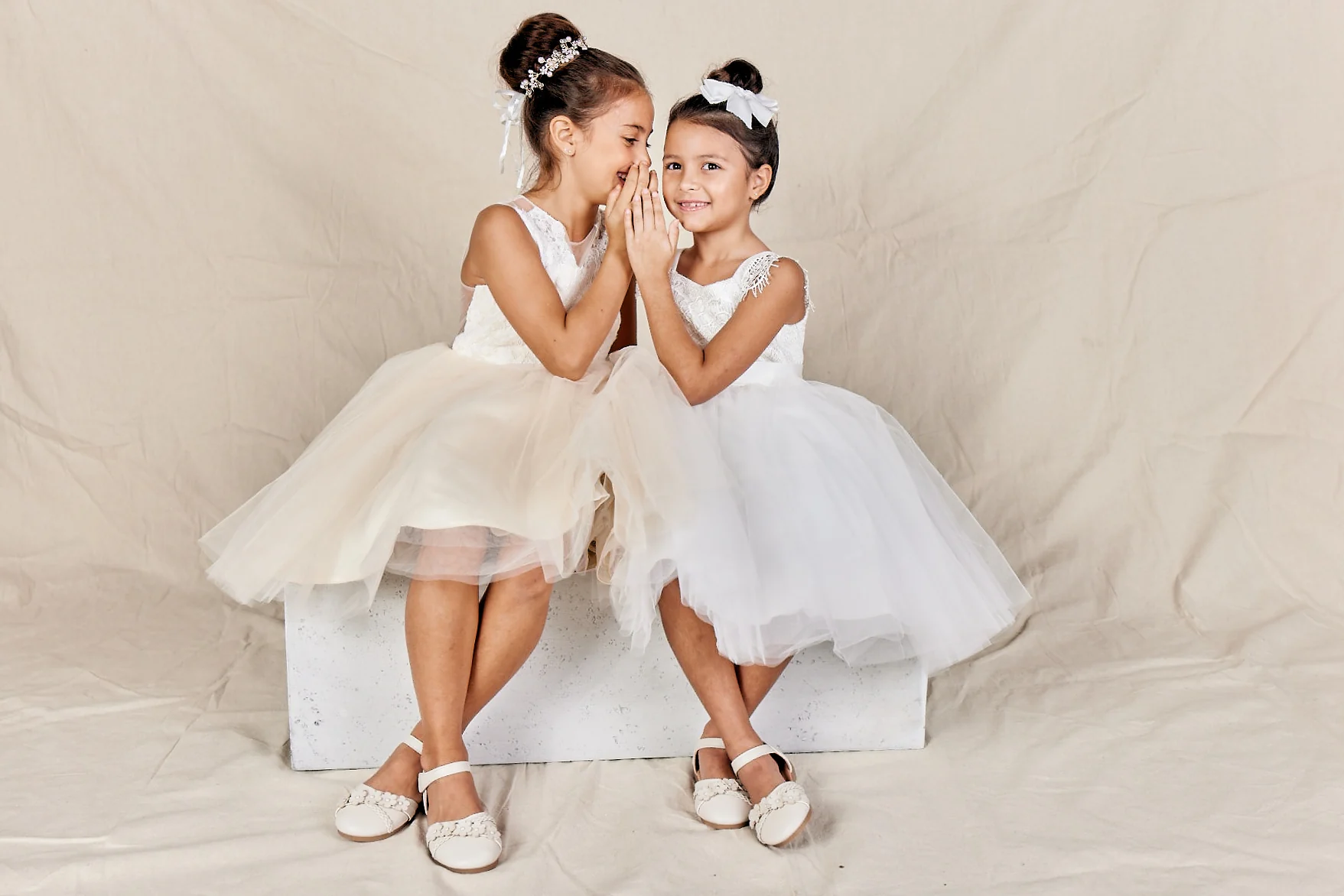 Two flower girls in ivory and white tulle dresses sharing a secret