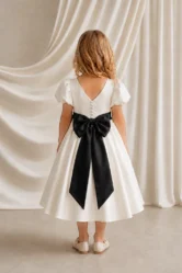 Back view of the Poppy ivory flower girl dress by TH&TH with a V-back, satin-covered buttons and an oversized black satin bow